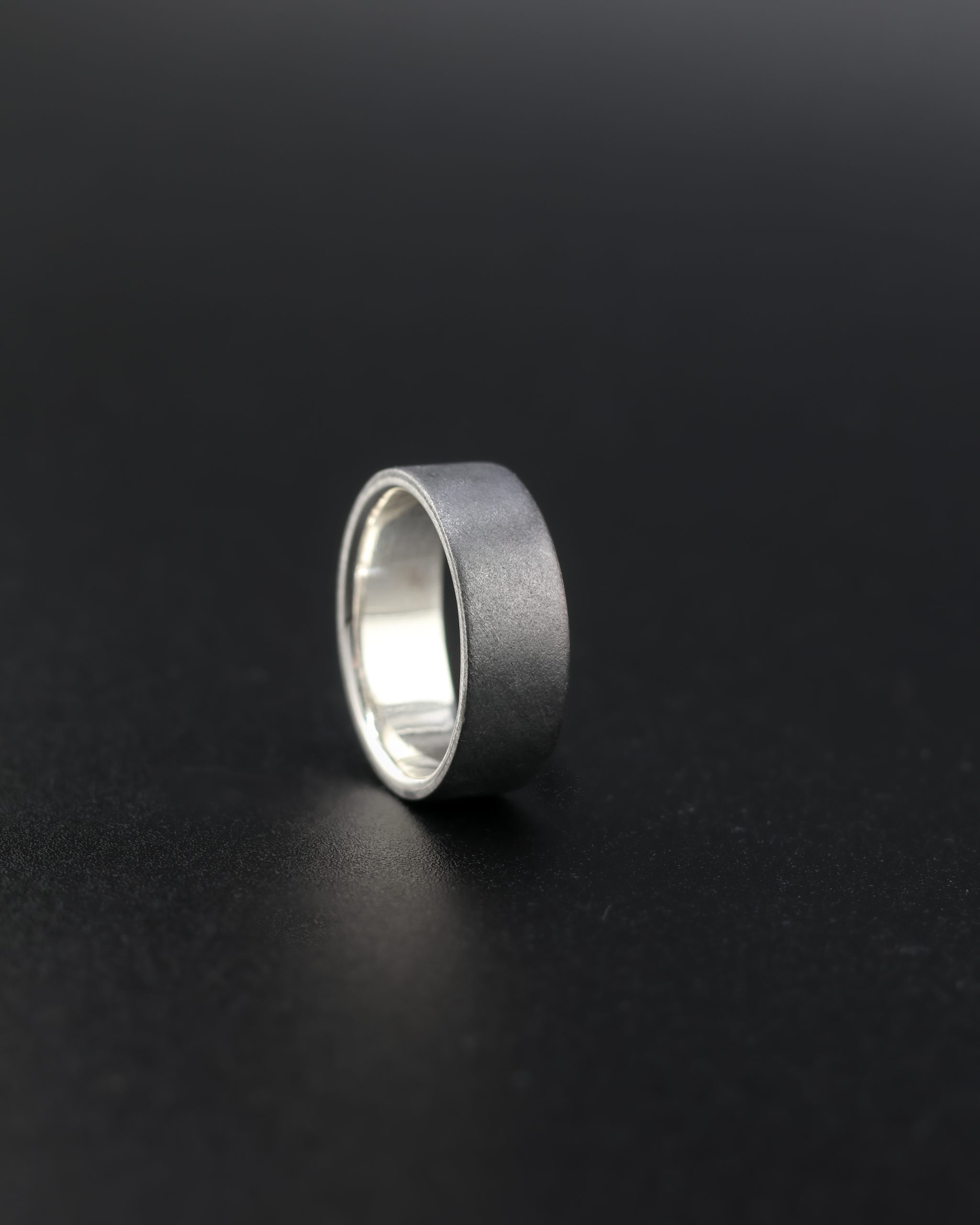 9E2A1943-2 Black Zirconium wedding ring which incorporates a sterling silver liner on the inside of the wedding ring. It has a low flat profile and is 7.5mm wide. Handmade Zirconium Rings in Devon, UK by award winning jeweller James French at JWF Unique Designs. This zirconium wedding ring is designed for someone seeking a durable, hardwearing ring which is also lightweight.