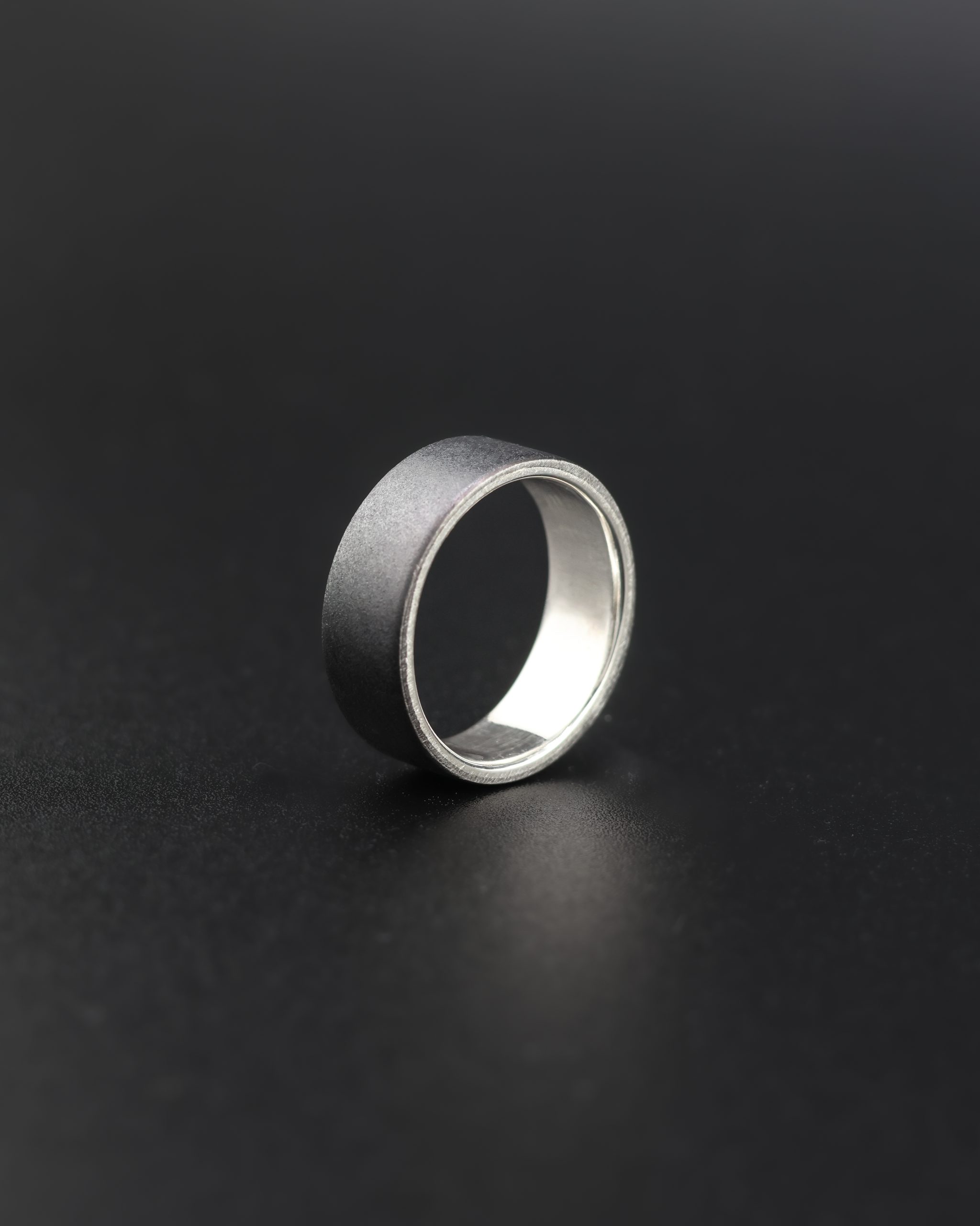 9E2A1944 Black Zirconium wedding ring which incorporates a sterling silver liner on the inside of the wedding ring. It has a low flat profile and is 7.5mm wide. Handmade Zirconium Rings in Devon, UK by award winning jeweller James French at JWF Unique Designs. This zirconium wedding ring is designed for someone seeking a durable, hardwearing ring which is also lightweight.