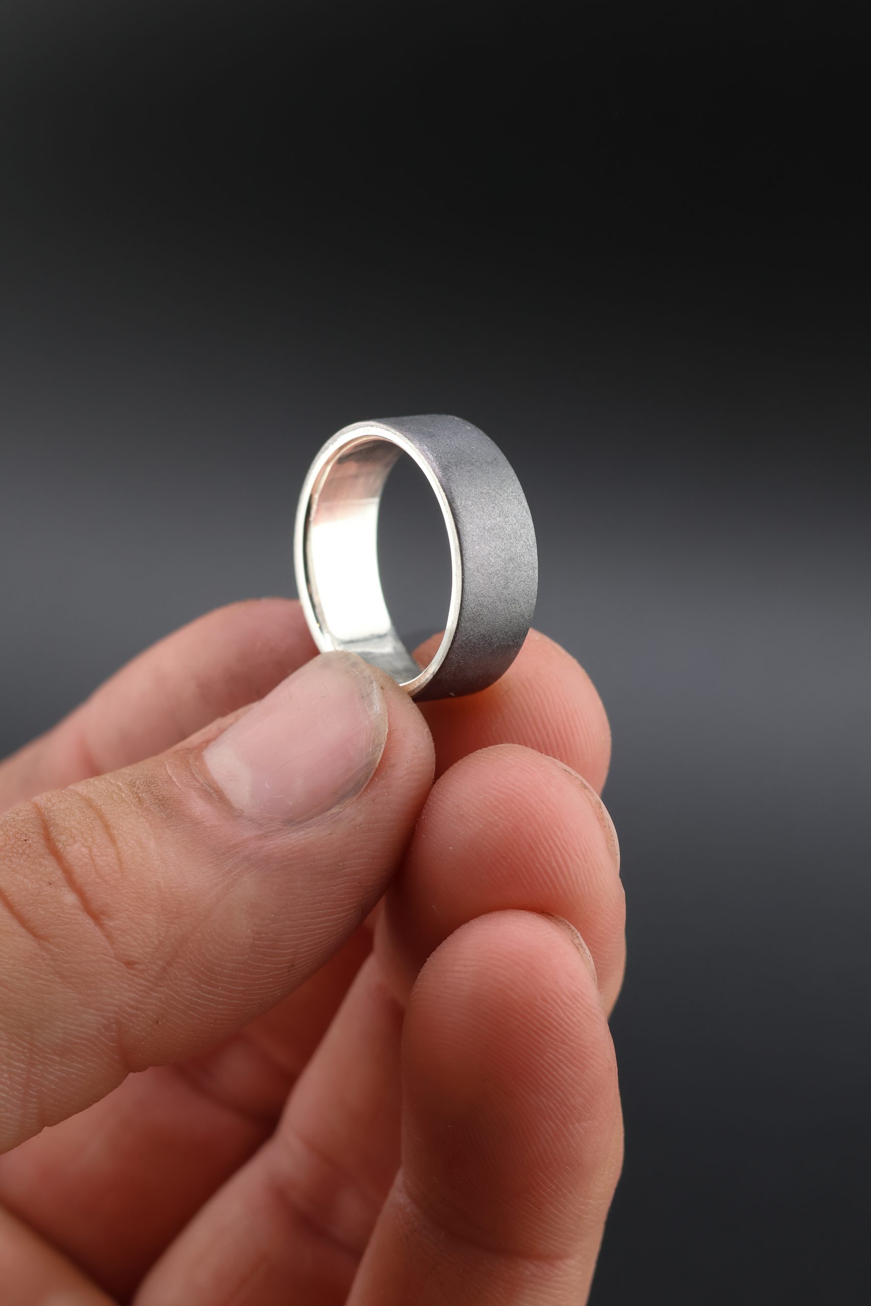 9E2A1958 Black Zirconium wedding ring which incorporates a sterling silver liner on the inside of the wedding ring. It has a low flat profile and is 7.5mm wide. Handmade Zirconium Rings in Devon, UK by award winning jeweller James French at JWF Unique Designs. This zirconium wedding ring is designed for someone seeking a durable, hardwearing ring which is also lightweight.