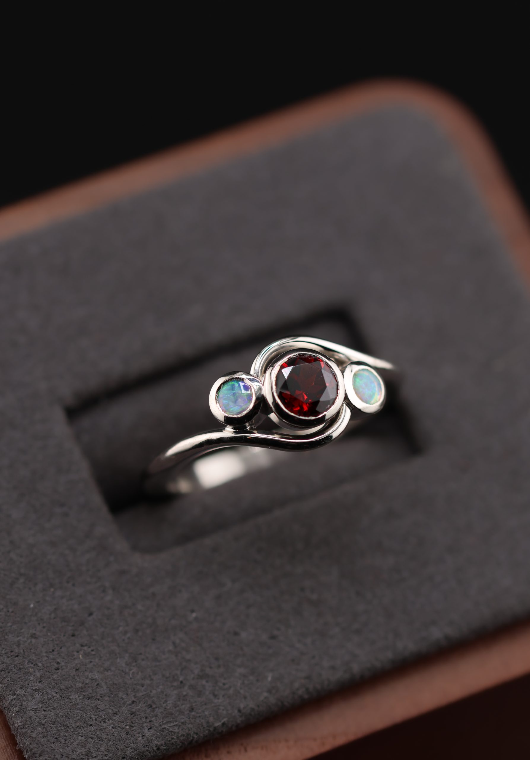 9E2A3580 Garnet and opal ladies ring with bezel set gemstones. Deep red garnet centre with two opals either side. High polish finish. Ideal for an engagement ring, milestone ring, or ladies gift. Handmade in Devon, UK. Made by award-winning goldsmith James French at JWF Unique Designs.