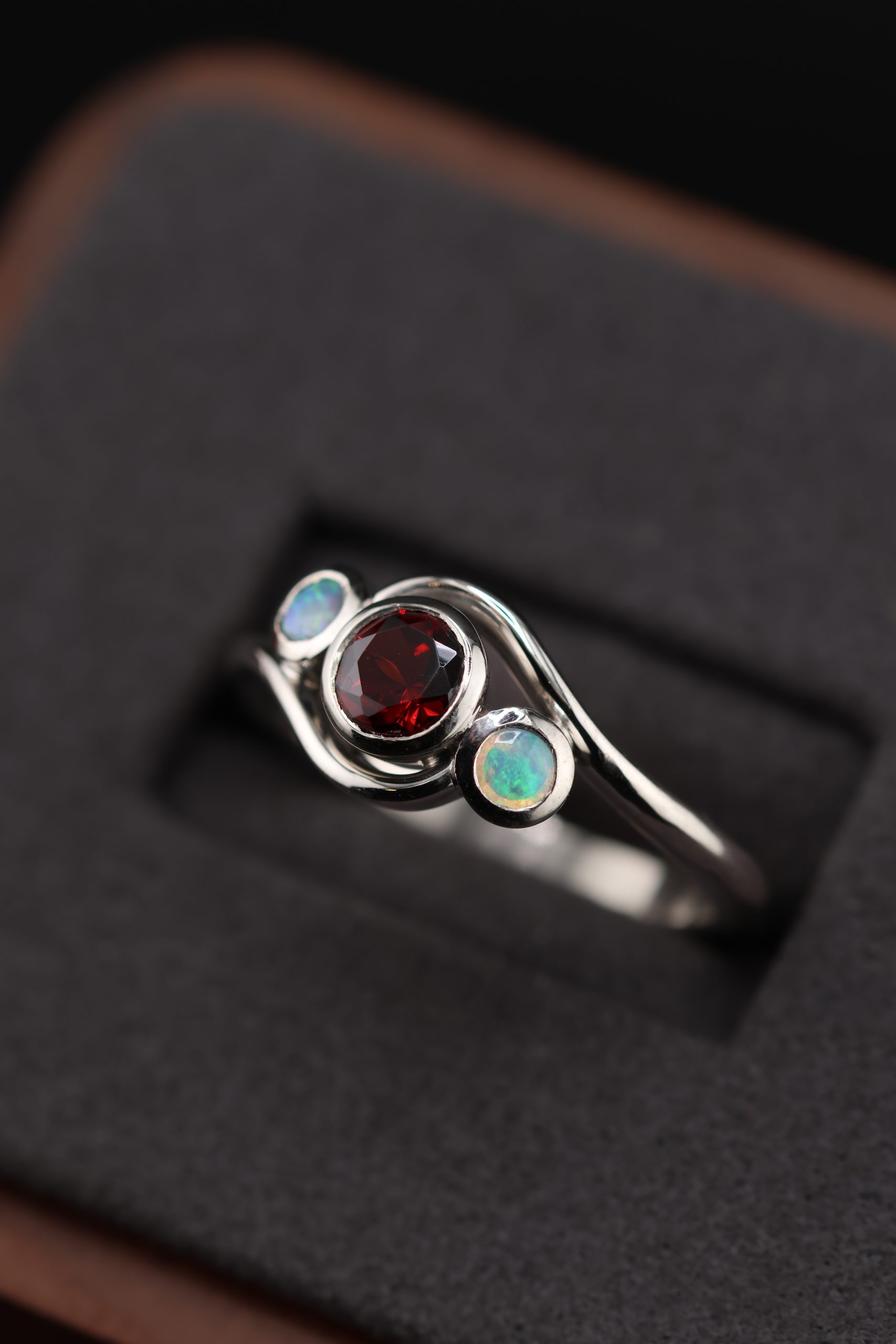9E2A3590 Garnet and opal ladies ring with bezel set gemstones. Deep red garnet centre with two opals either side. High polish finish. Ideal for an engagement ring, milestone ring, or ladies gift. Handmade in Devon, UK. Made by award-winning goldsmith James French at JWF Unique Designs.