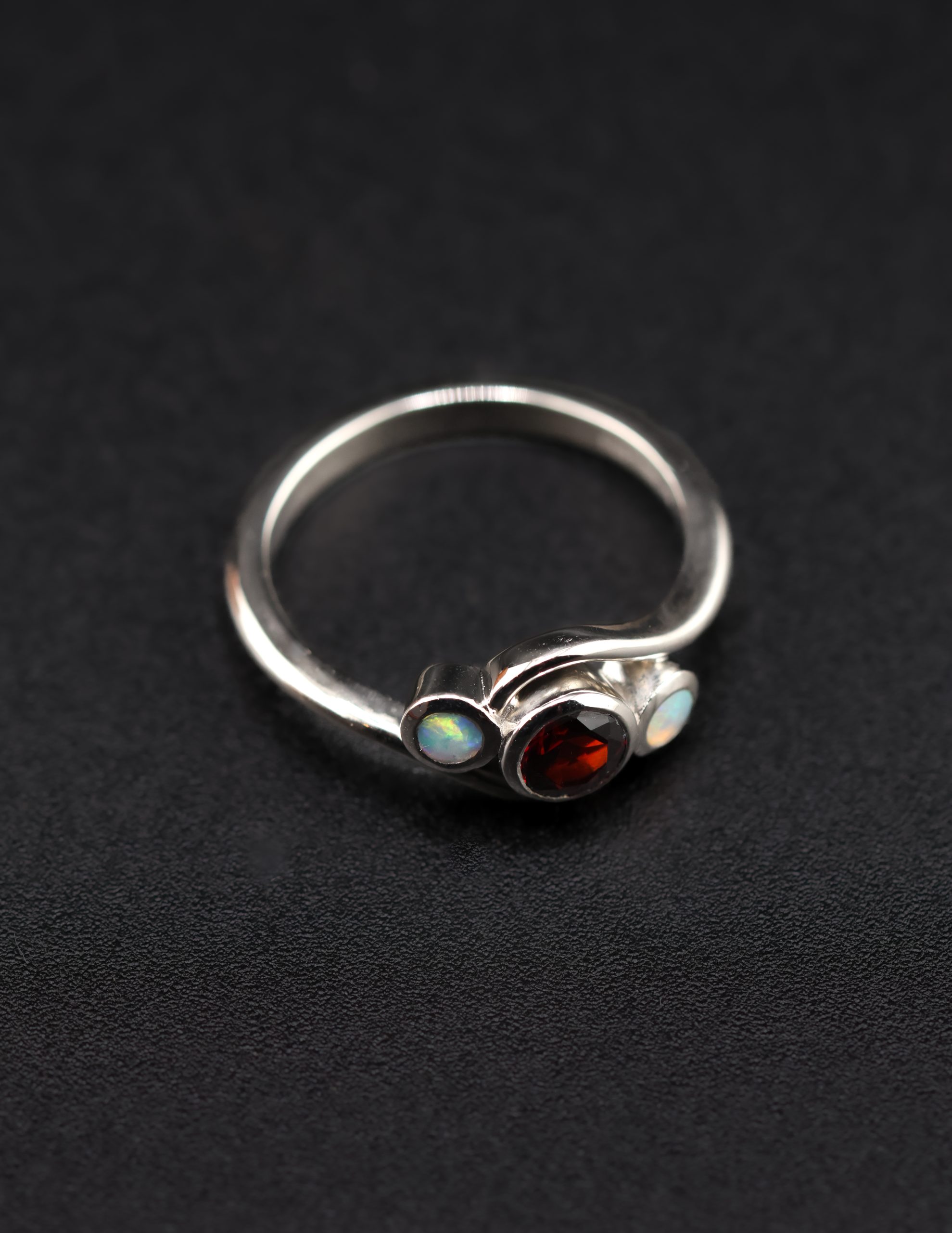9E2A3596 Garnet and opal ladies ring with bezel set gemstones. Deep red garnet centre with two opals either side. High polish finish. Ideal for an engagement ring, milestone ring, or ladies gift. Handmade in Devon, UK. Made by award-winning goldsmith James French at JWF Unique Designs.