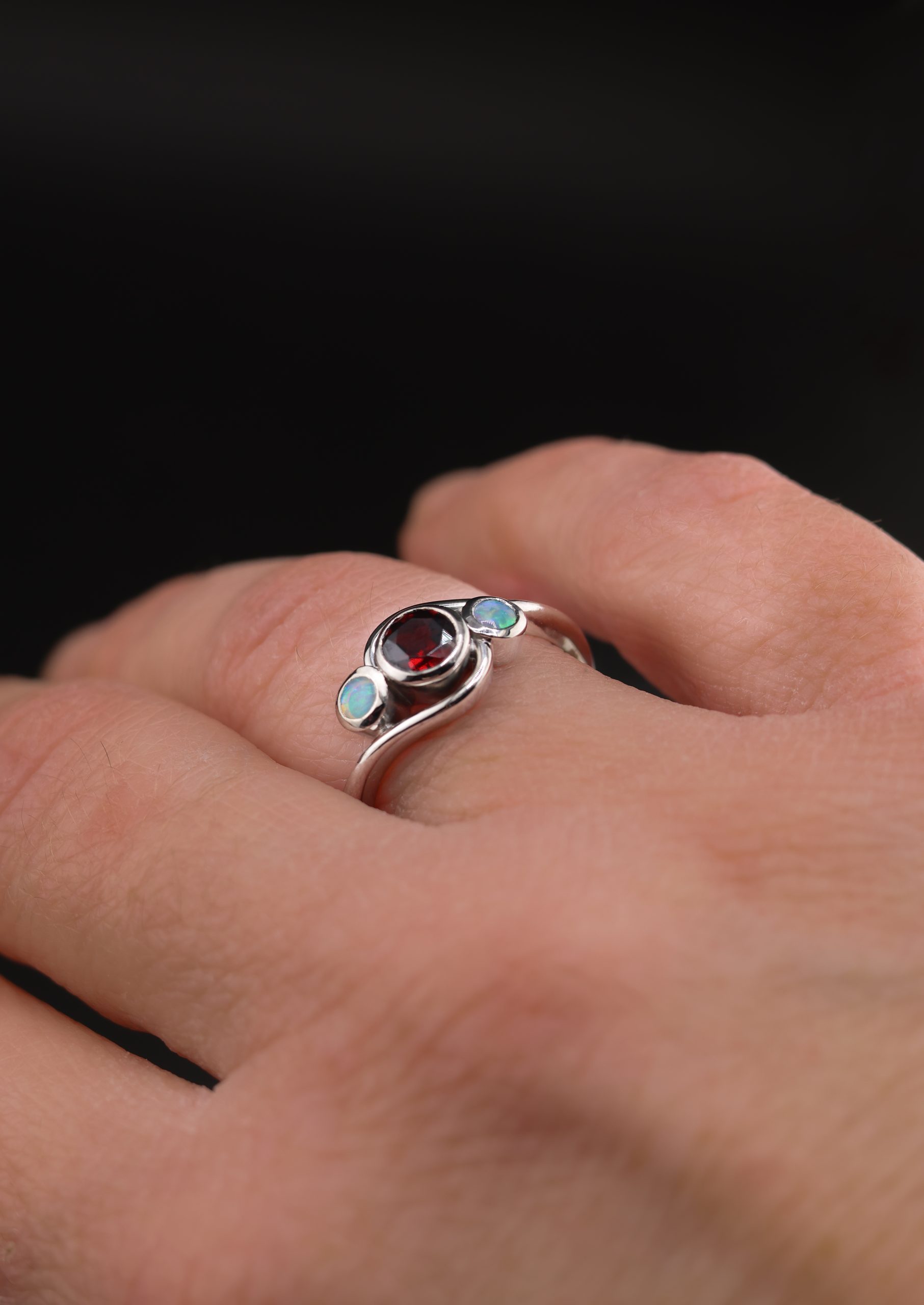 9E2A3599 Garnet and opal ladies ring with bezel set gemstones. Deep red garnet centre with two opals either side. High polish finish. Ideal for an engagement ring, milestone ring, or ladies gift. Handmade in Devon, UK. Made by award-winning goldsmith James French at JWF Unique Designs.