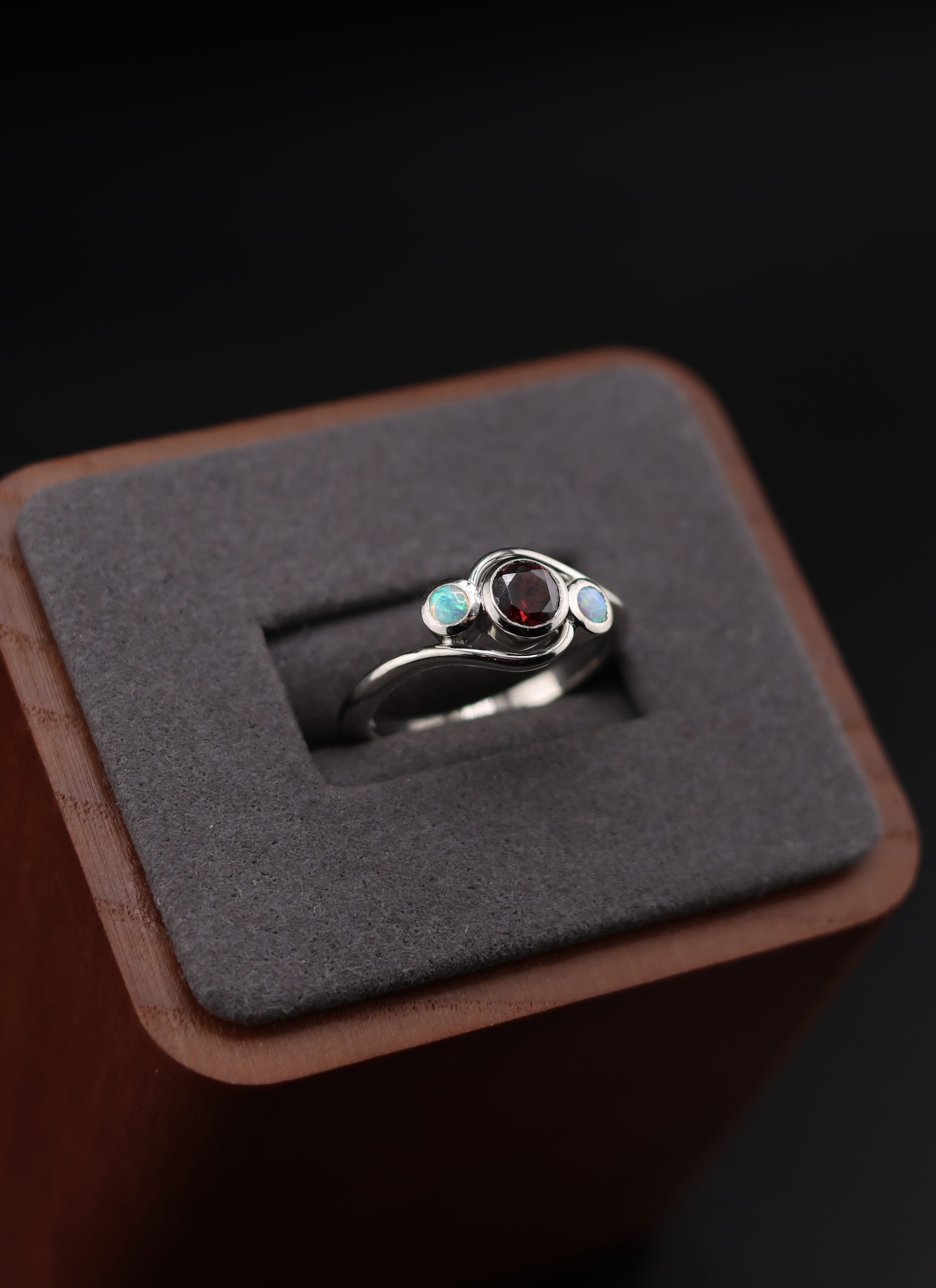 9E2A3606 Garnet and opal ladies ring with bezel set gemstones. Deep red garnet centre with two opals either side. High polish finish. Ideal for an engagement ring, milestone ring, or ladies gift. Handmade in Devon, UK. Made by award-winning goldsmith James French at JWF Unique Designs.