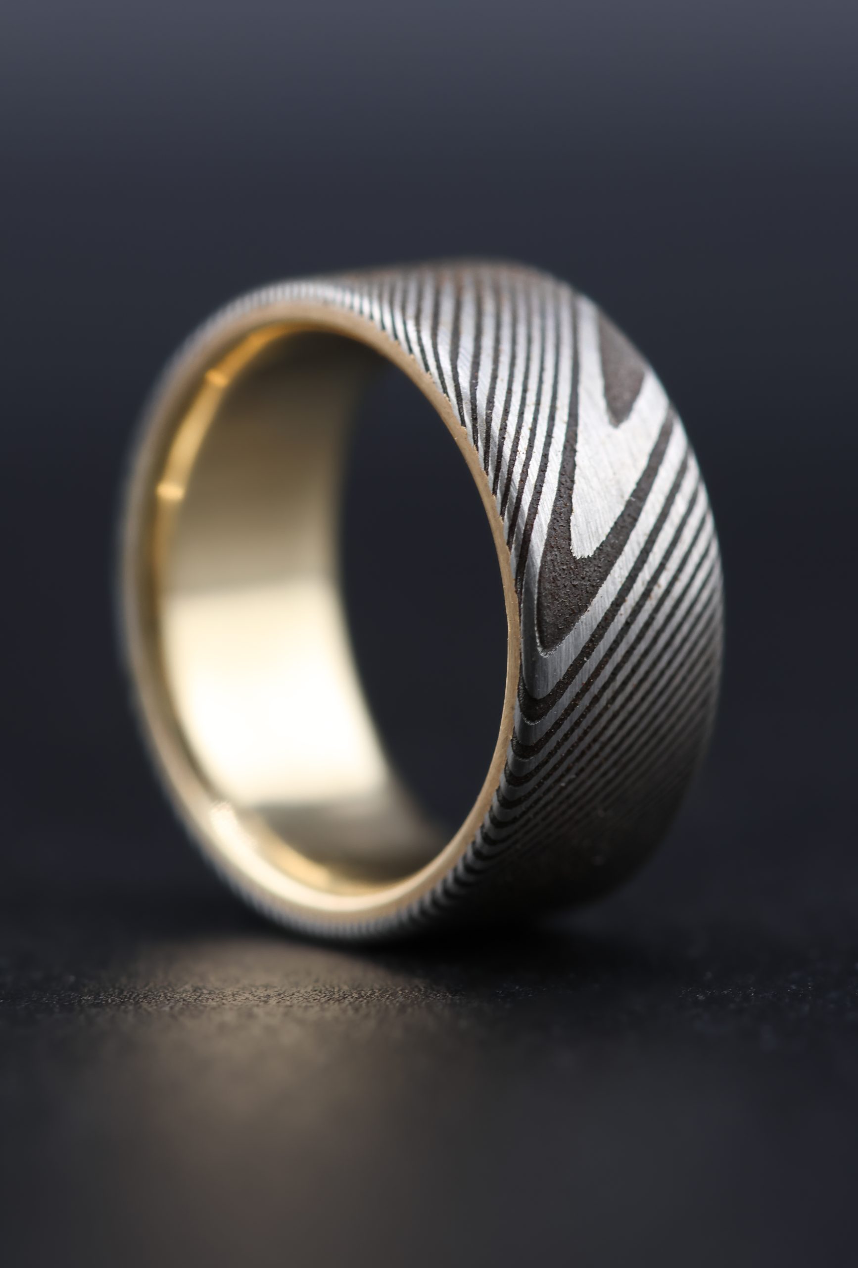 9E2A4171 Damascus steel wedding ring with a yellow gold liner. Twisted pattern throughout the band with darker laminations. Flat profile with original stainless steel colour. Available with a 9ct or 18ct yellow gold liner. Lightweight, durable and maintenance free men's wedding ring. Handmade in Devon, UK by award-winning goldsmith James French.