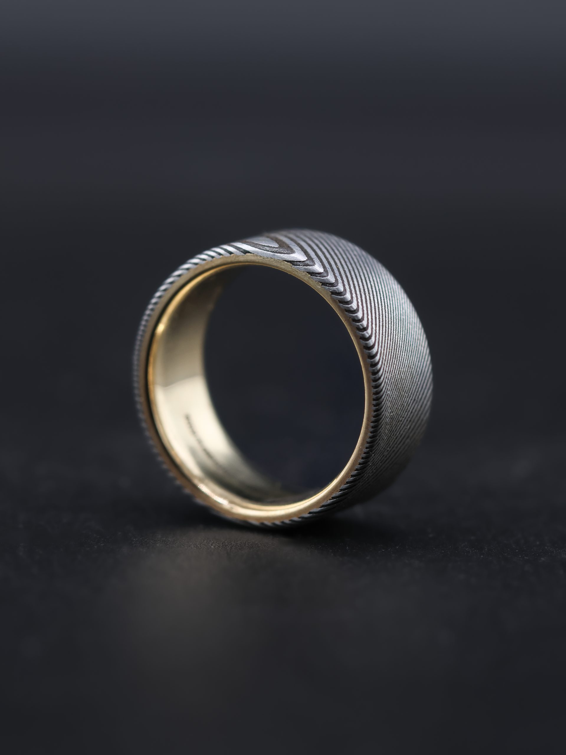 9E2A4177 Damascus steel wedding ring with a yellow gold liner. Twisted pattern throughout the band with darker laminations. Flat profile with original stainless steel colour. Available with a 9ct or 18ct yellow gold liner. Lightweight, durable and maintenance free men's wedding ring. Handmade in Devon, UK by award-winning goldsmith James French.