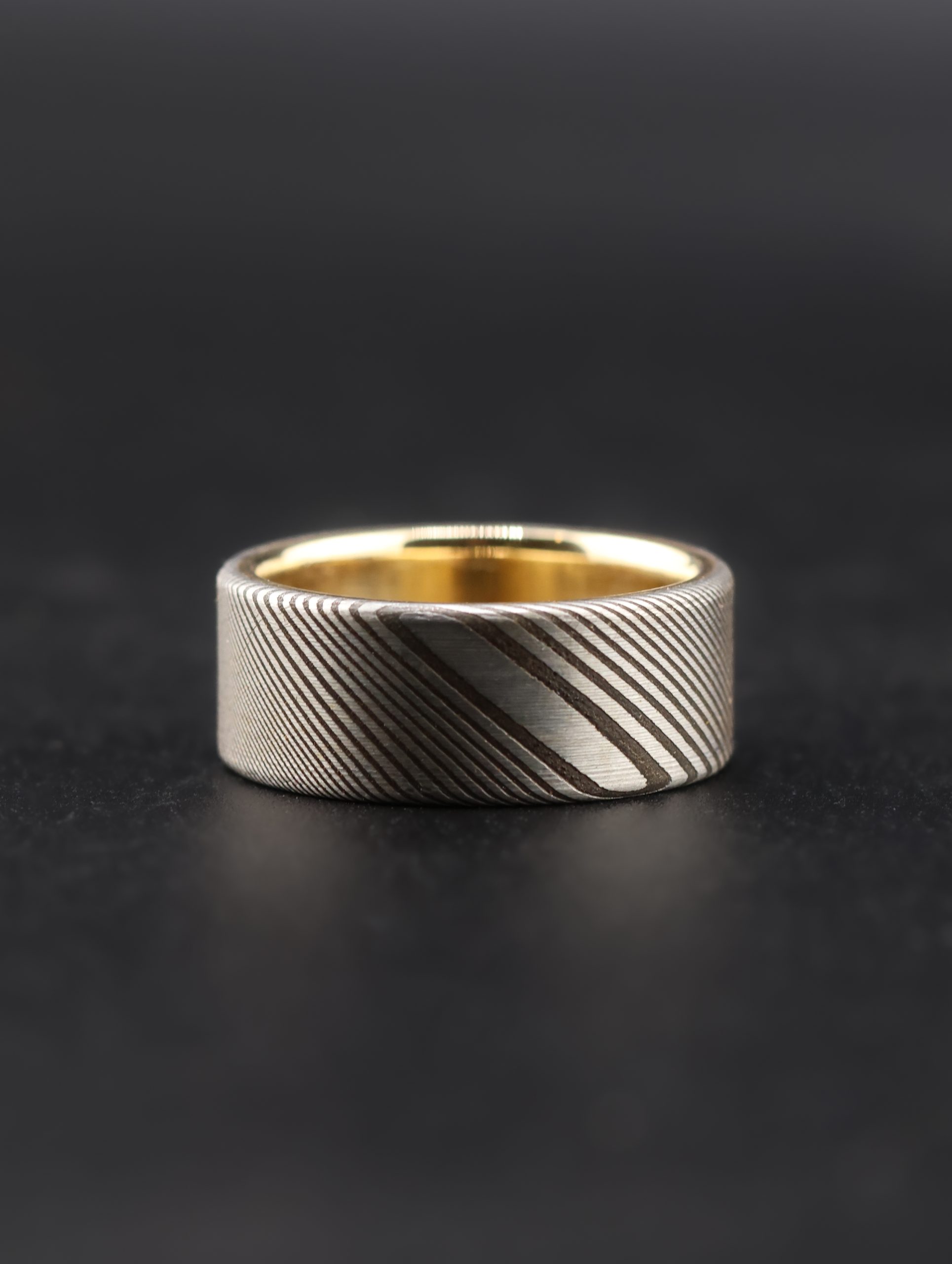 9E2A4179 Damascus steel wedding ring with a yellow gold liner. Twisted pattern throughout the band with darker laminations. Flat profile with original stainless steel colour. Available with a 9ct or 18ct yellow gold liner. Lightweight, durable and maintenance free men's wedding ring. Handmade in Devon, UK by award-winning goldsmith James French.