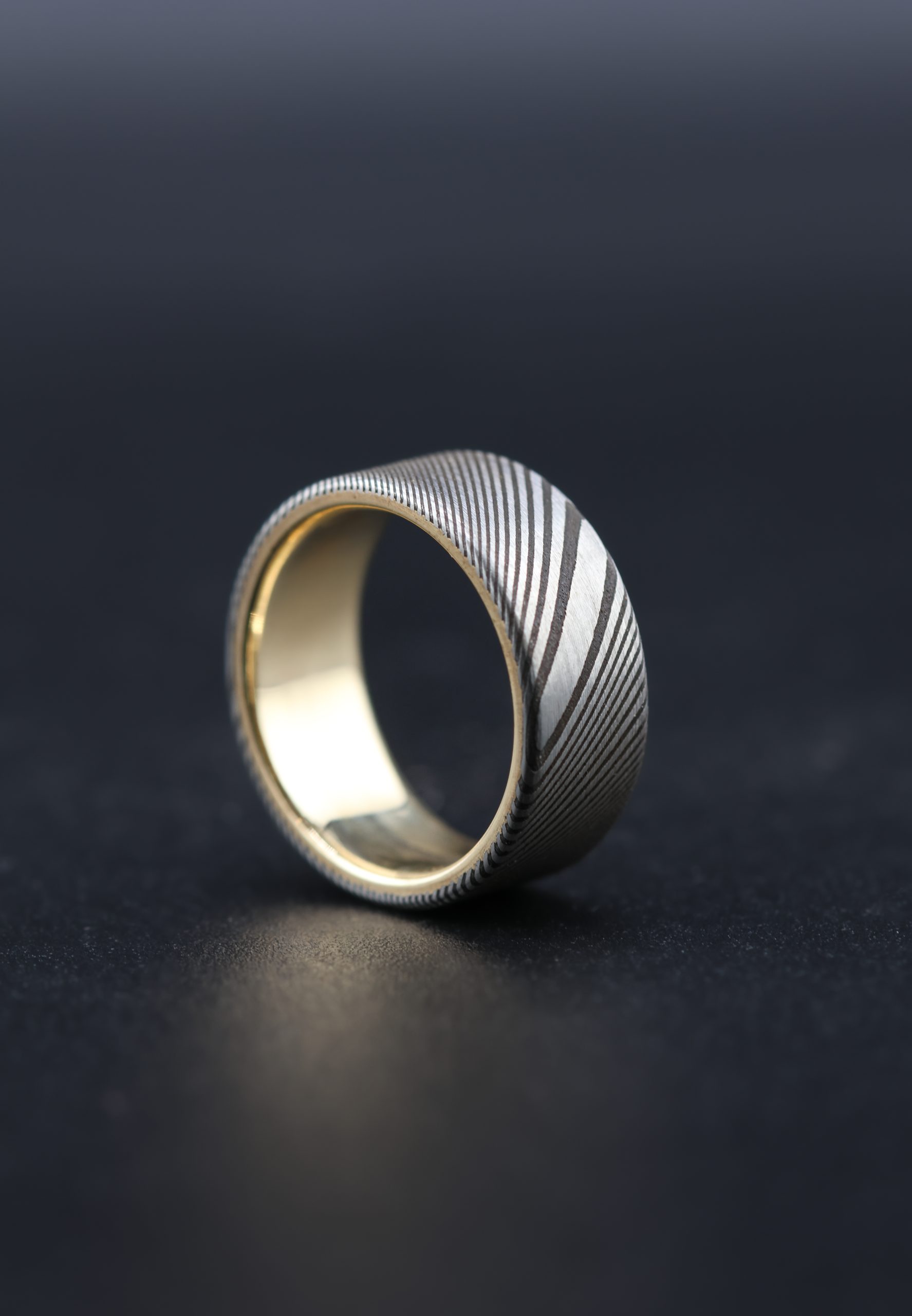 9E2A4180 Damascus steel wedding ring with a yellow gold liner. Twisted pattern throughout the band with darker laminations. Flat profile with original stainless steel colour. Available with a 9ct or 18ct yellow gold liner. Lightweight, durable and maintenance free men's wedding ring. Handmade in Devon, UK by award-winning goldsmith James French.