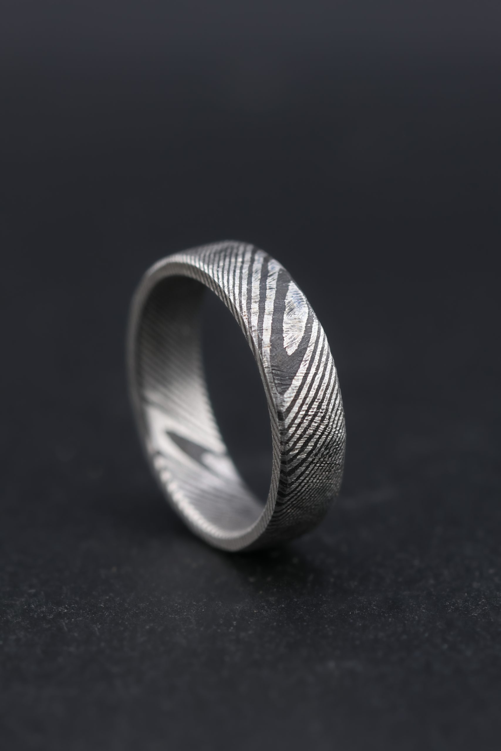 9E2A4181 Damascus steel wedding ring with a twisted pattern and cast finish. Original silver tone steel with darker layered laminations. Slightly rounded profile with a unique pattern throughout the band. Lightweight, durable and maintenance free men's wedding ring. Handmade in Devon, UK by award-winning goldsmith James French.