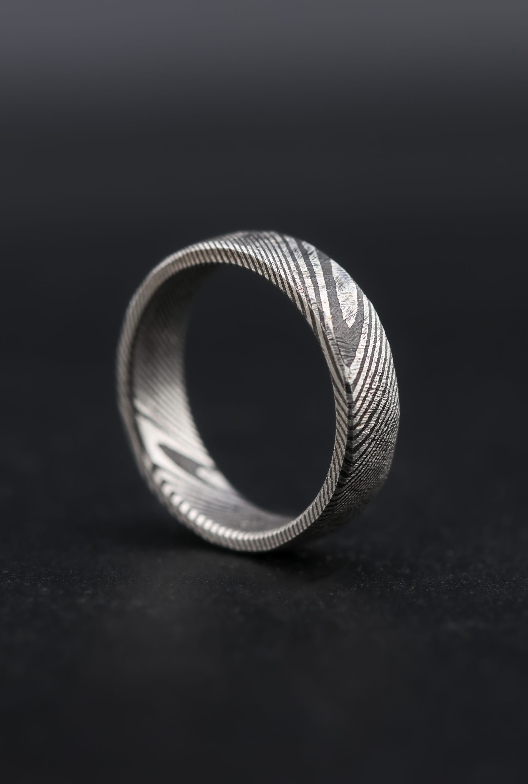 9E2A4182-3 Damascus steel wedding ring with a twisted pattern and cast finish. Original silver tone steel with darker layered laminations. Slightly rounded profile with a unique pattern throughout the band. Lightweight, durable and maintenance free men's wedding ring. Handmade in Devon, UK by award-winning goldsmith James French.