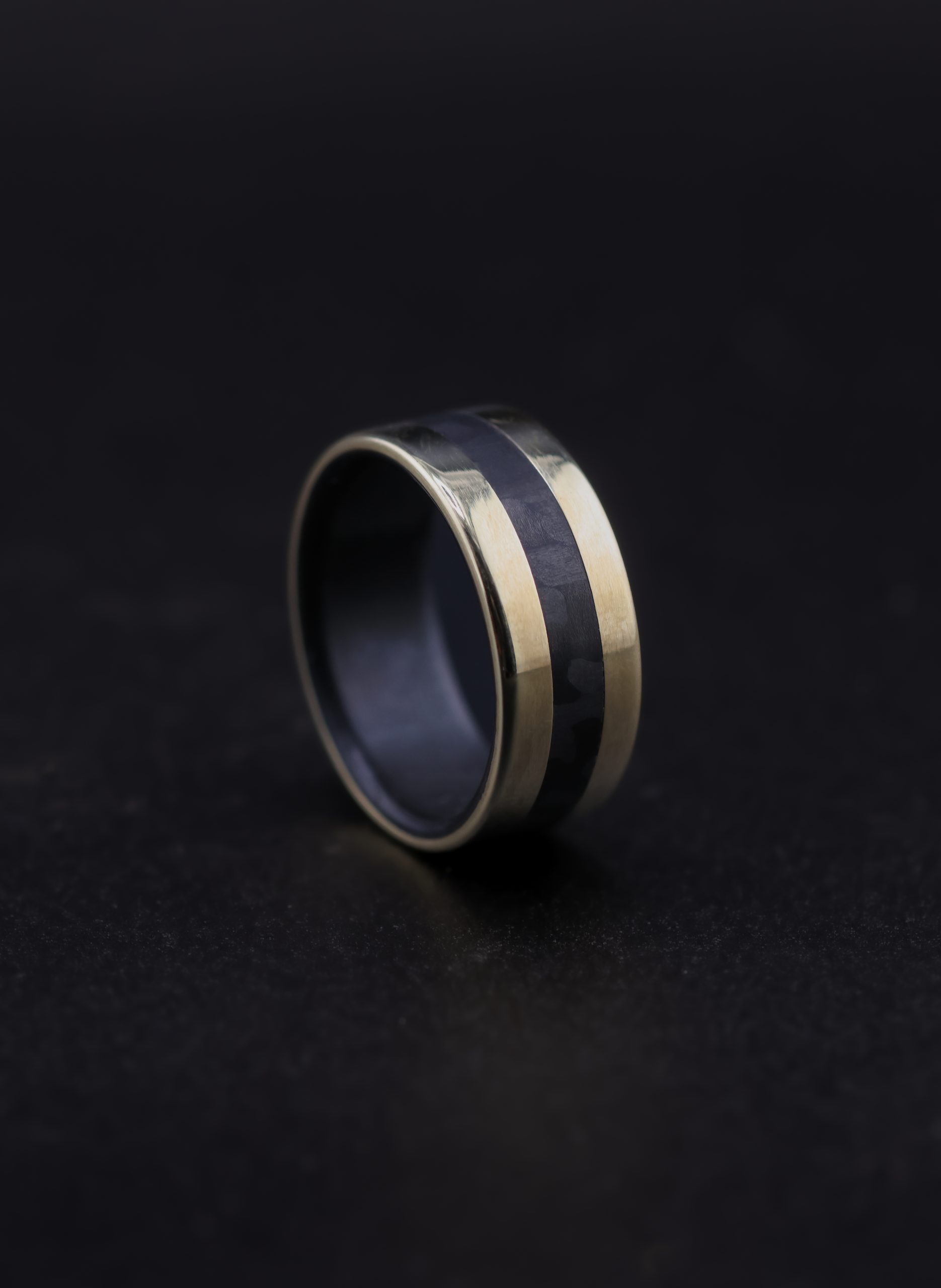 9E2A5668-2 9ct yellow gold wedding ring with black carbon fibre inlay and liner. Flat profile band with polished finish. 3mm carbon fibre centre inlay. 9mm wide men's wedding ring. Handmade in Devon, UK by award-winning goldsmith James French.