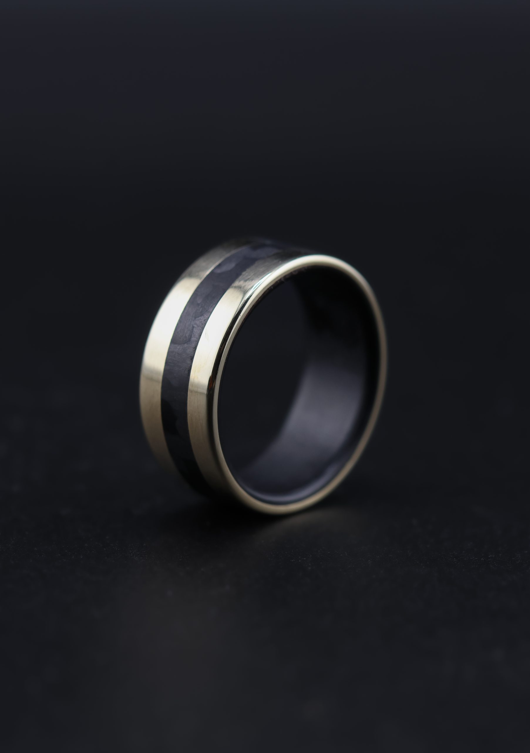 9E2A5673 9ct yellow gold wedding ring with black carbon fibre inlay and liner. Flat profile band with polished finish. 3mm carbon fibre centre inlay. 9mm wide men's wedding ring. Handmade in Devon, UK by award-winning goldsmith James French.