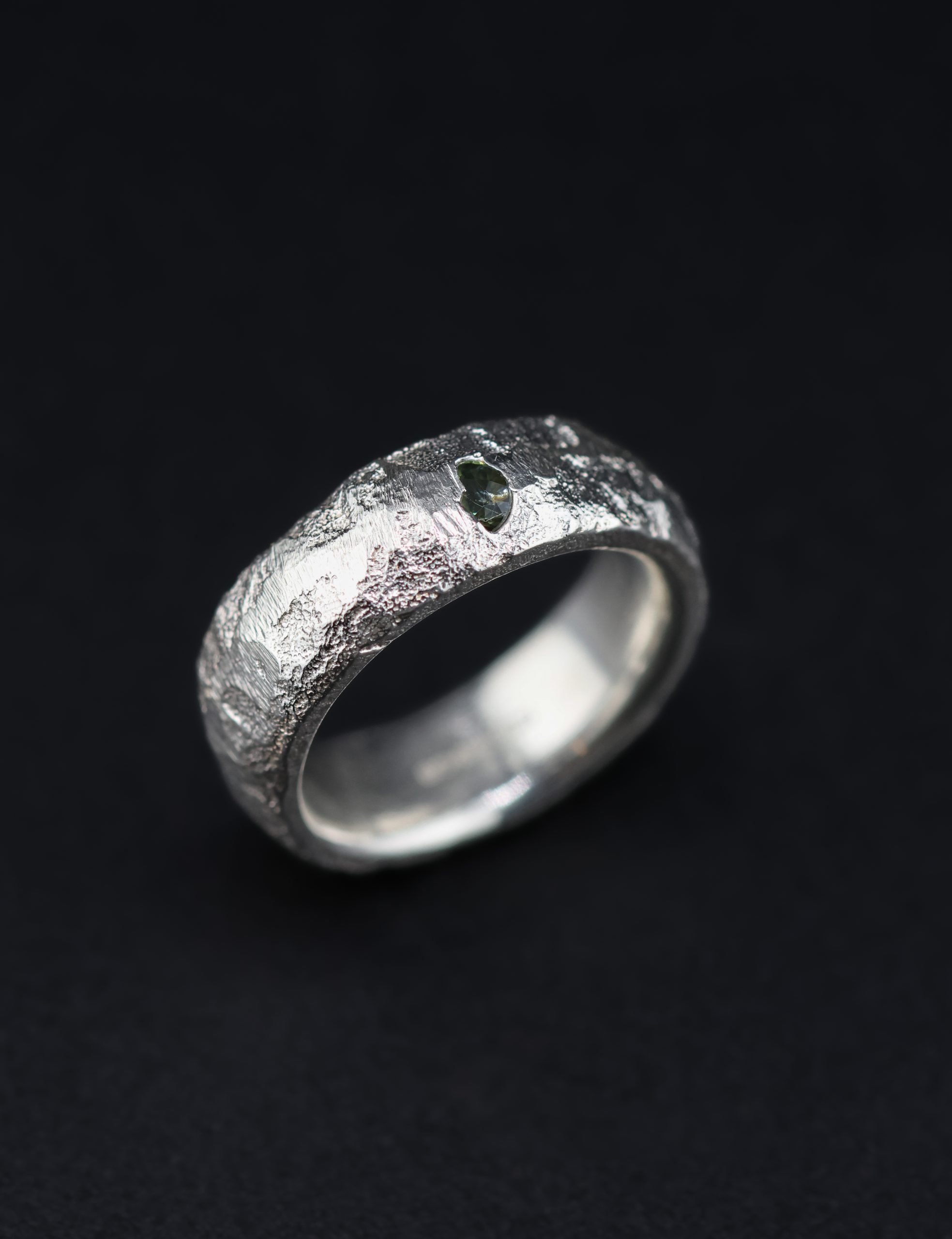 9E2A5749 A 7mm sterling silver sand cast wedding ring with a D-shaped profile and rugged organic texture. A green sapphire sits directly within the uneven silver surface. This distinctive gemstone wedding ring is handmade in Devon by goldsmith James French.