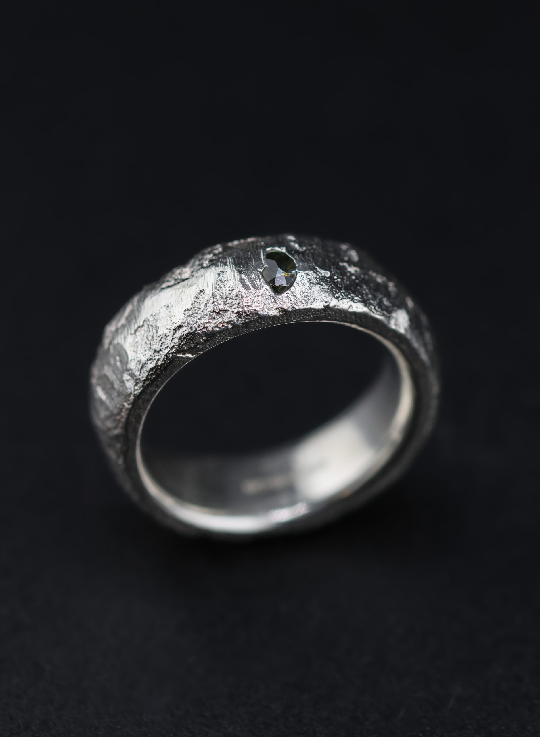 9E2A5751 A 7mm sterling silver sand cast wedding ring with a D-shaped profile and rugged organic texture. A green sapphire sits directly within the uneven silver surface. This distinctive gemstone wedding ring is handmade in Devon by goldsmith James French.