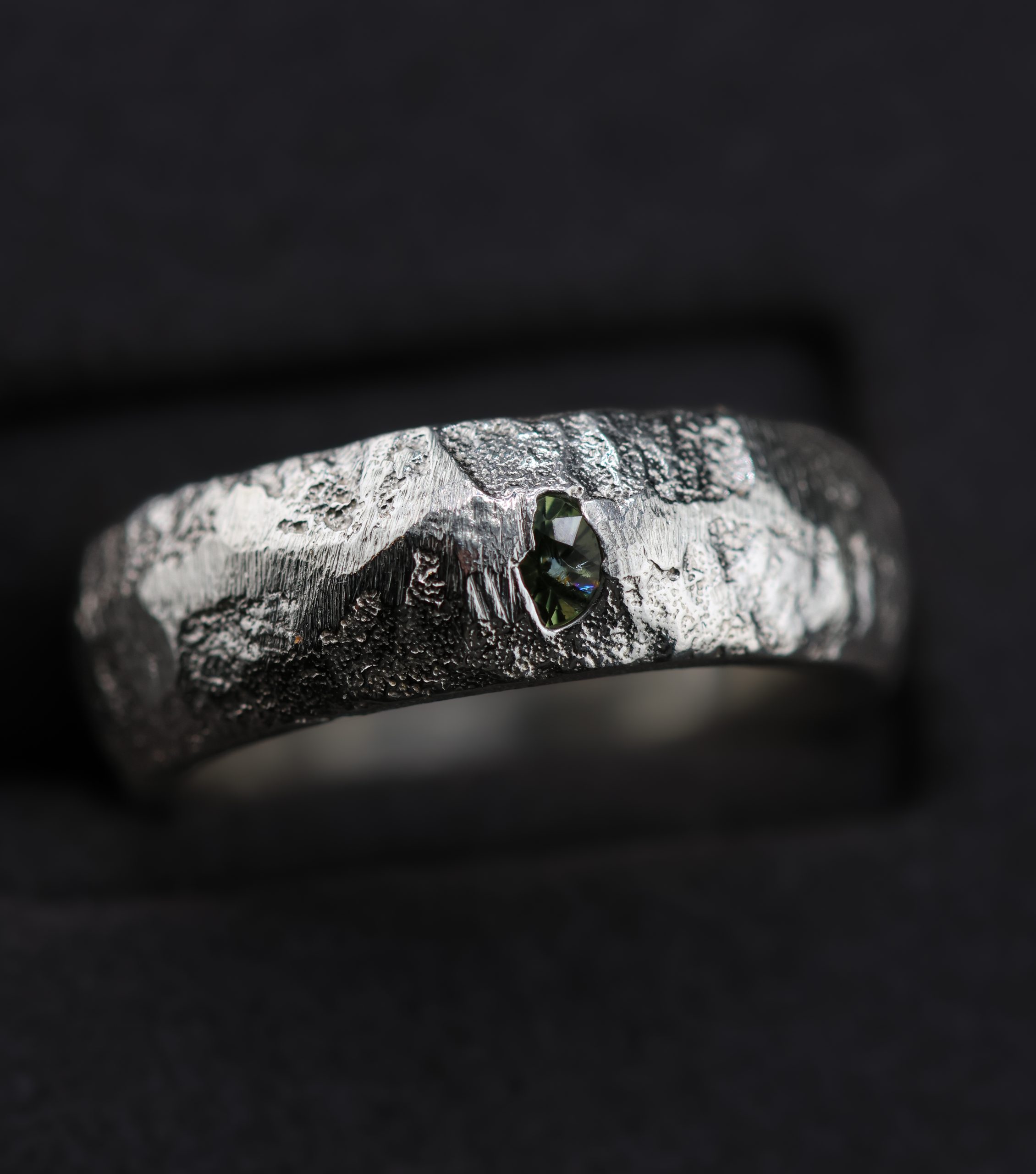 9E2A5756 A 7mm sterling silver sand cast wedding ring with a D-shaped profile and rugged organic texture. A green sapphire sits directly within the uneven silver surface. This distinctive gemstone wedding ring is handmade in Devon by goldsmith James French.