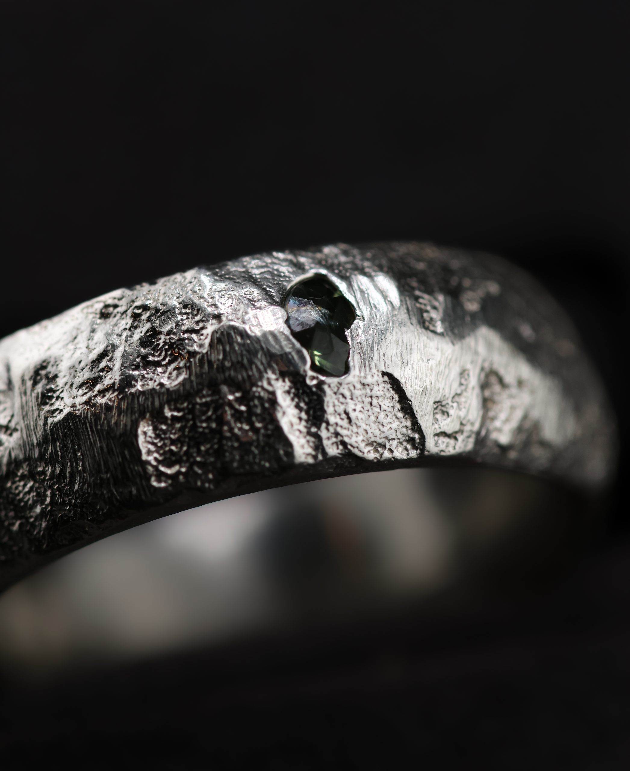 9E2A5758 A 7mm sterling silver sand cast wedding ring with a D-shaped profile and rugged organic texture. A green sapphire sits directly within the uneven silver surface. This distinctive gemstone wedding ring is handmade in Devon by goldsmith James French.