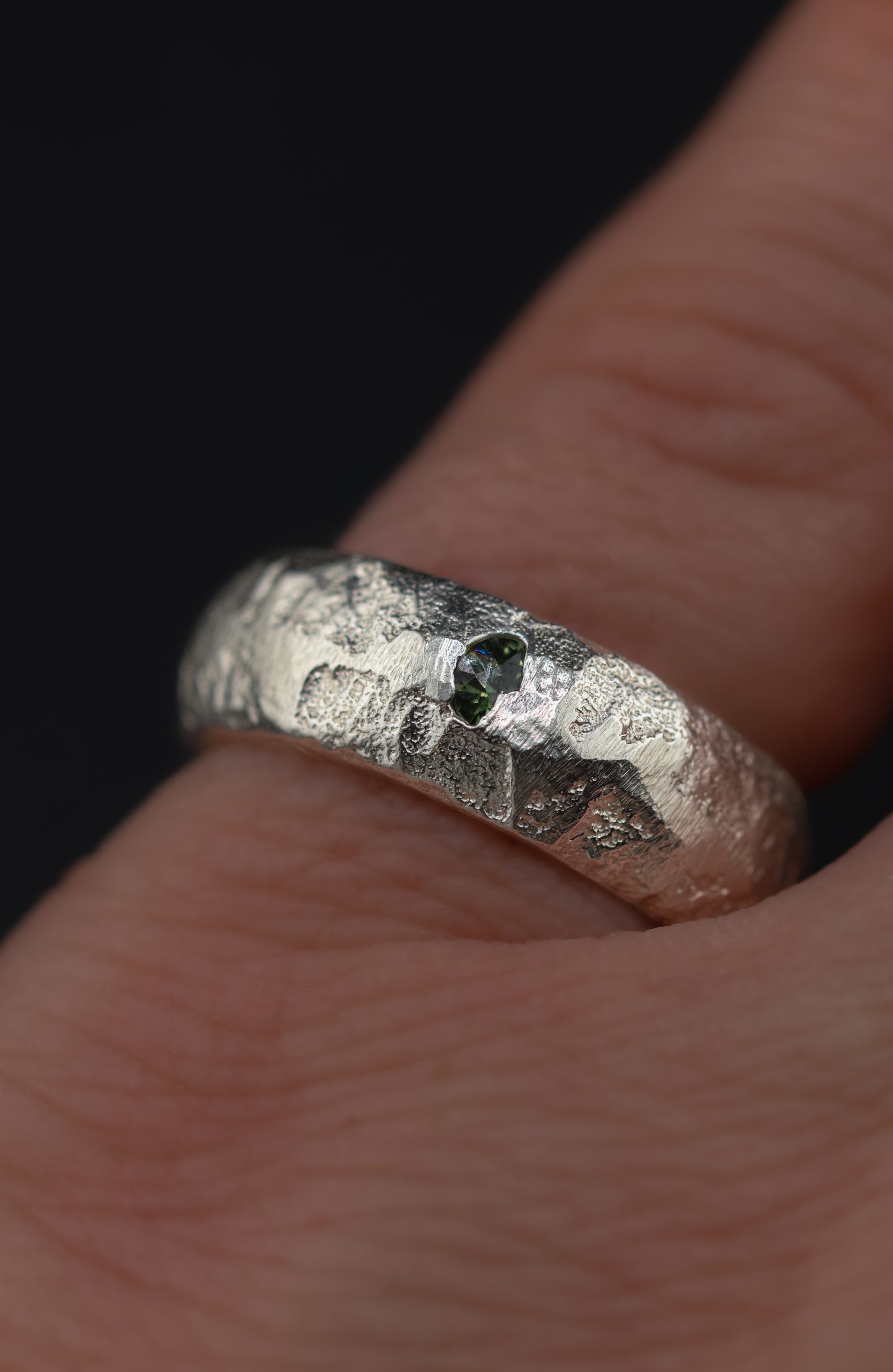 9E2A5762 A 7mm sterling silver sand cast wedding ring with a D-shaped profile and rugged organic texture. A green sapphire sits directly within the uneven silver surface. This distinctive gemstone wedding ring is handmade in Devon by goldsmith James French.