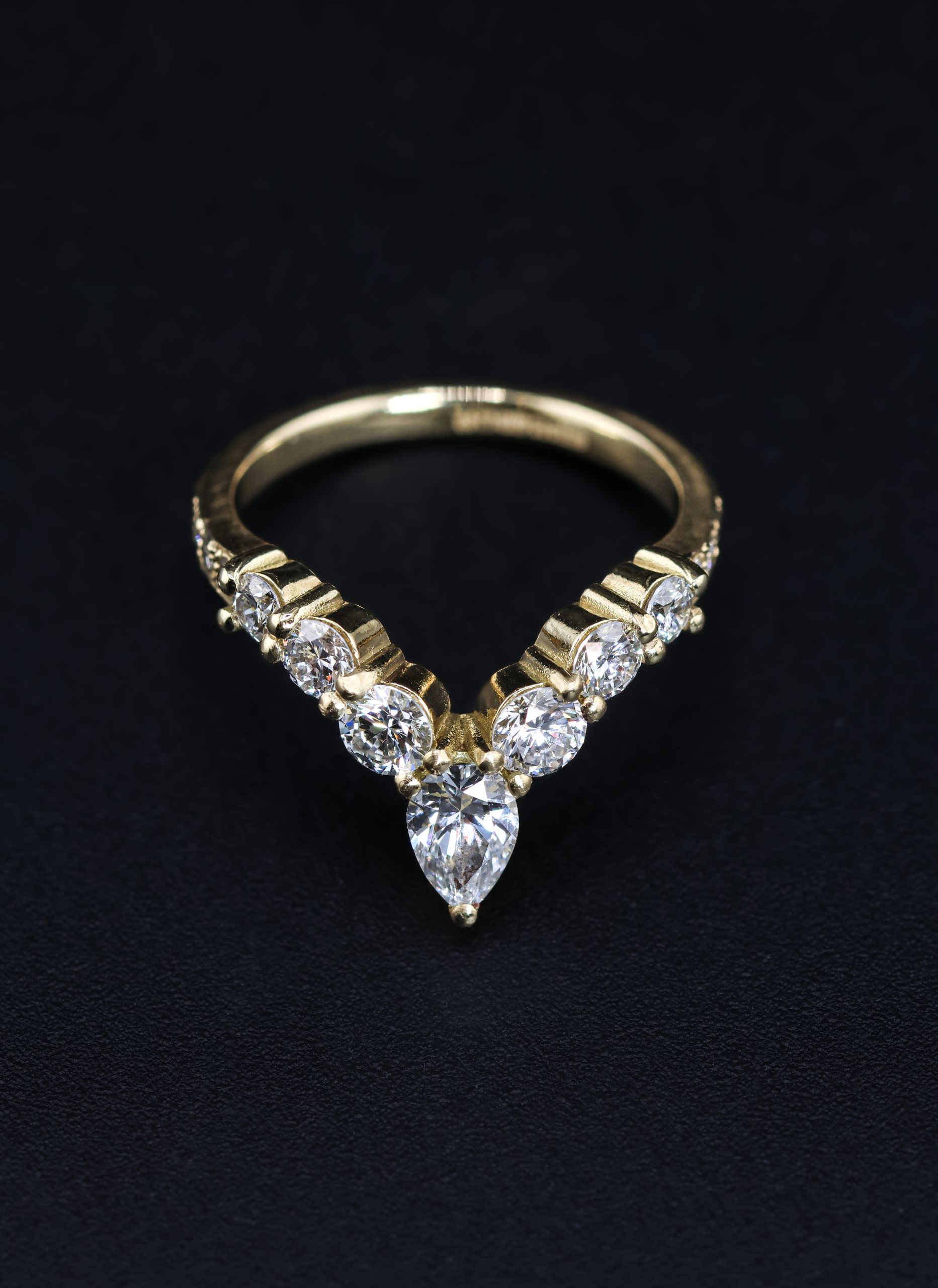 9E2A5824 An 18ct yellow gold shaped wedding ring designed to fit around an engagement ring. The swept design features a central pear-shaped diamond with round natural and lab-grown diamonds flowing along the curved shank. This bespoke diamond wedding ring has a high-polished finish. Handmade in Devon by goldsmith James French.