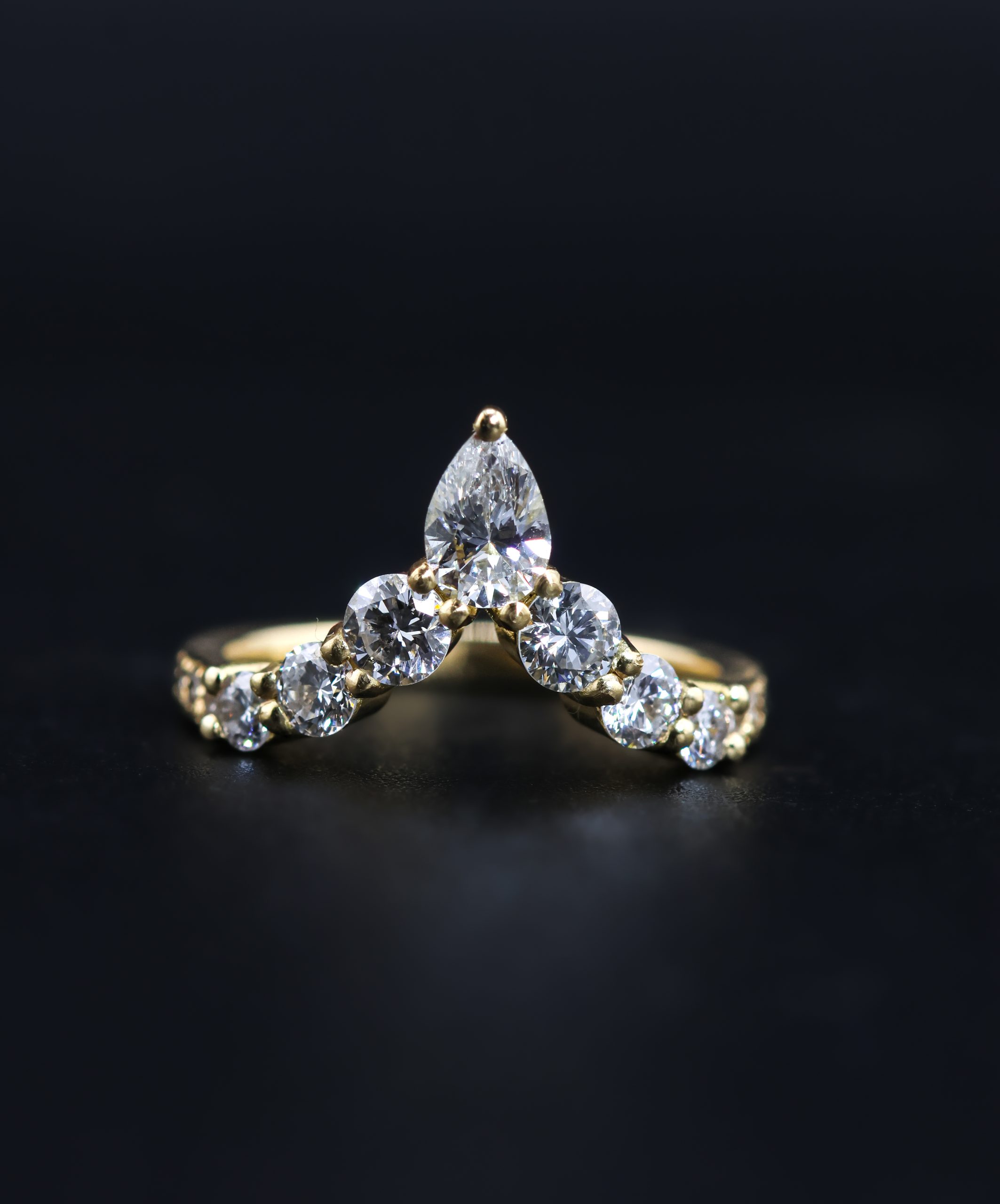9E2A5829 An 18ct yellow gold shaped wedding ring designed to fit around an engagement ring. The swept design features a central pear-shaped diamond with round natural and lab-grown diamonds flowing along the curved shank. This bespoke diamond wedding ring has a high-polished finish. Handmade in Devon by goldsmith James French.