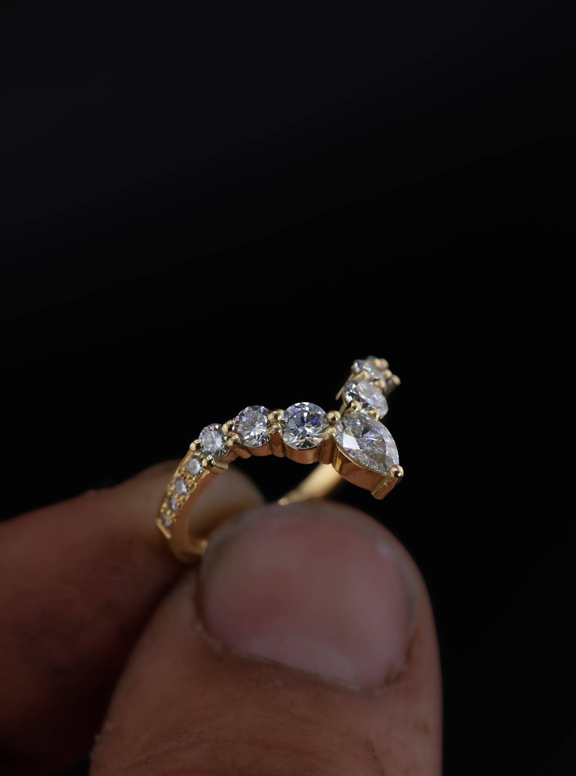 9E2A5839 An 18ct yellow gold shaped wedding ring designed to fit around an engagement ring. The swept design features a central pear-shaped diamond with round natural and lab-grown diamonds flowing along the curved shank. This bespoke diamond wedding ring has a high-polished finish. Handmade in Devon by goldsmith James French.