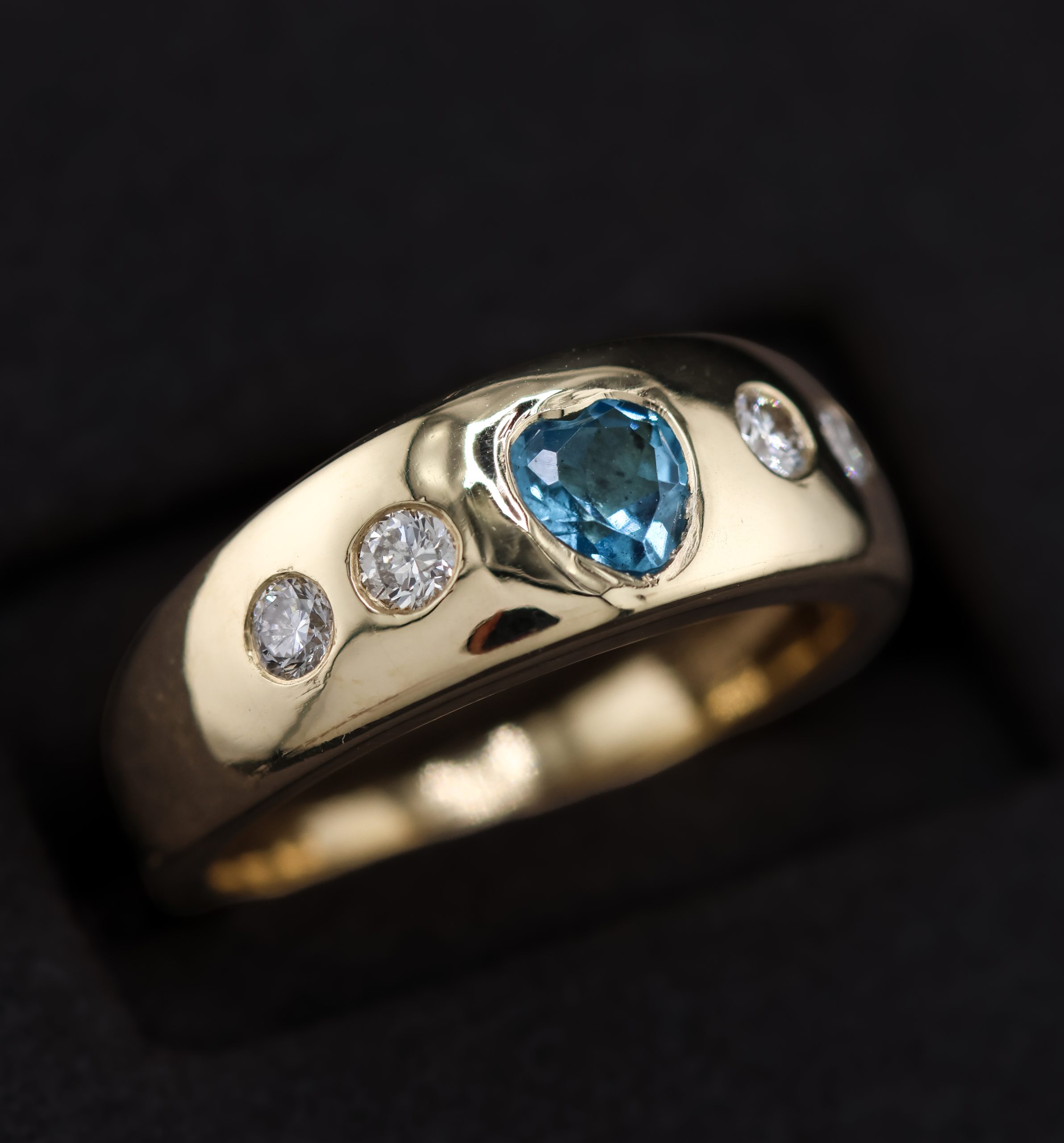 9E2A5863 A wide yellow gold ring featuring a heart-shaped blue topaz with two round diamonds on each side. The polished ring is handmade in Devon by goldsmith and silversmith James French.