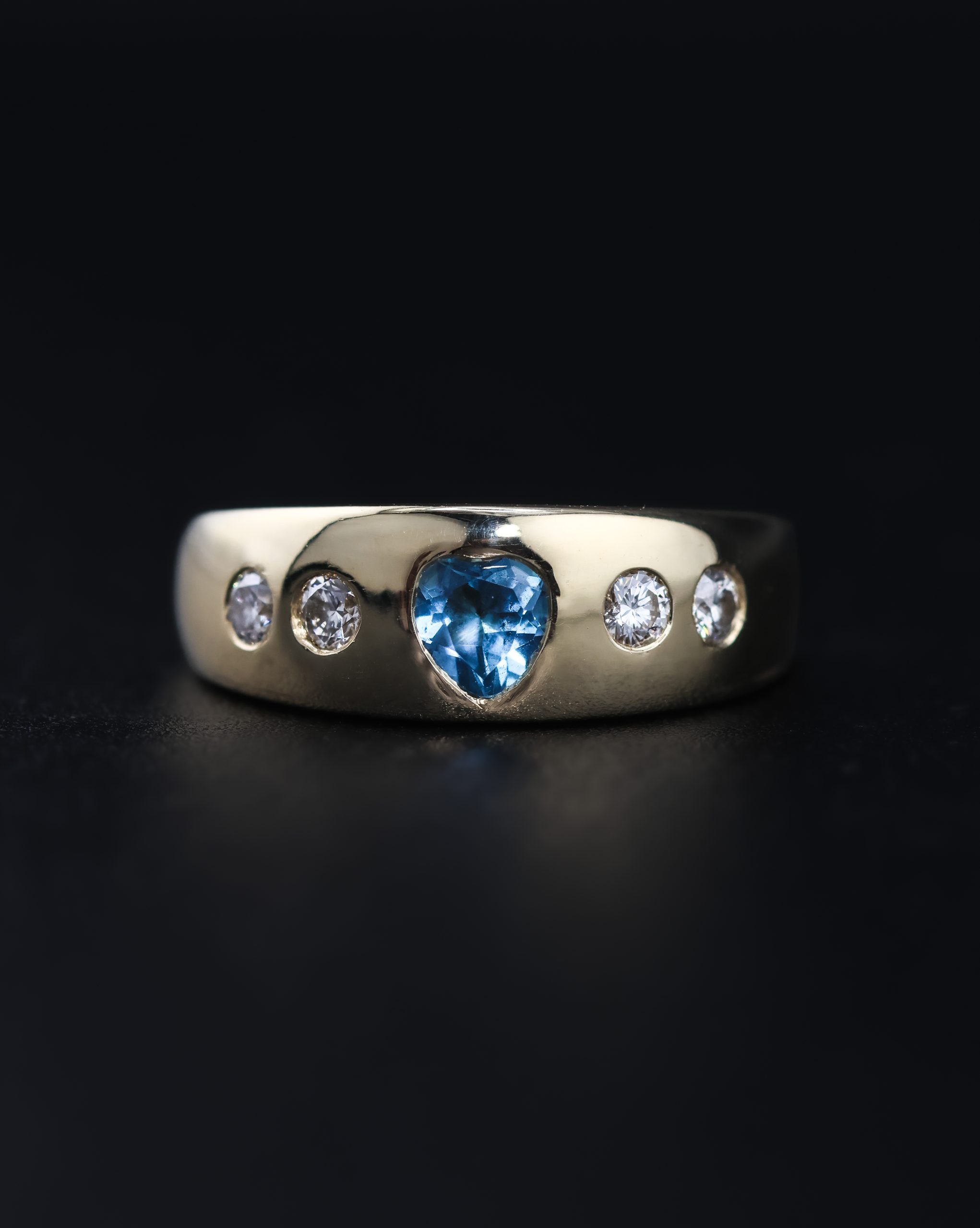 9E2A5867 A wide yellow gold ring featuring a heart-shaped blue topaz with two round diamonds on each side. The polished ring is handmade in Devon by goldsmith and silversmith James French.