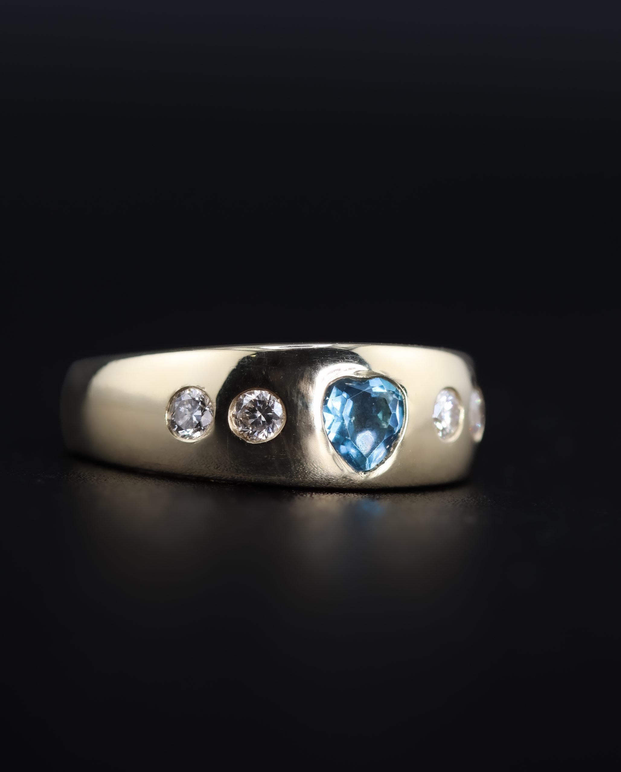 9E2A5870 A wide yellow gold ring featuring a heart-shaped blue topaz with two round diamonds on each side. The polished ring is handmade in Devon by goldsmith and silversmith James French.