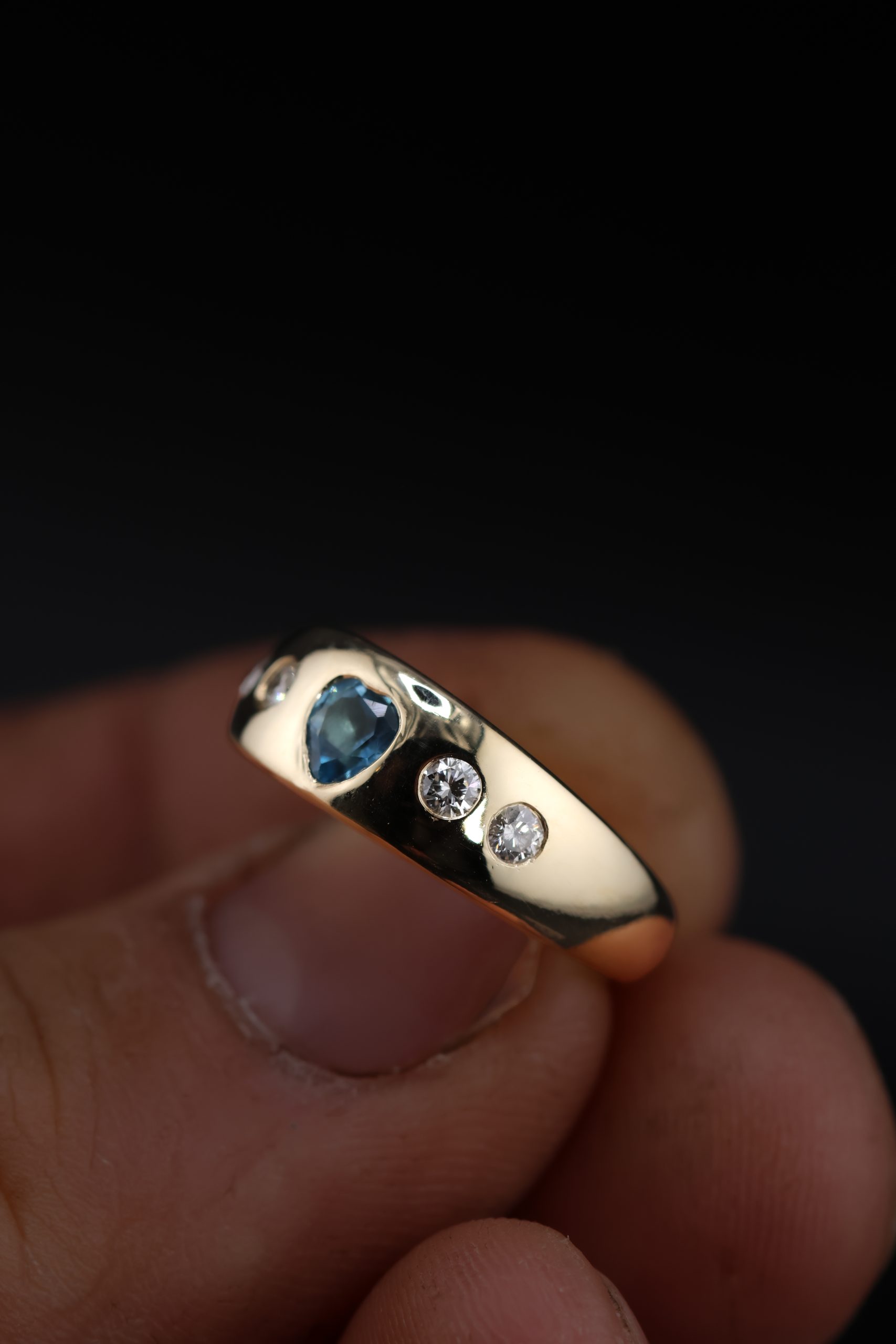9E2A5873 A wide yellow gold ring featuring a heart-shaped blue topaz with two round diamonds on each side. The polished ring is handmade in Devon by goldsmith and silversmith James French.